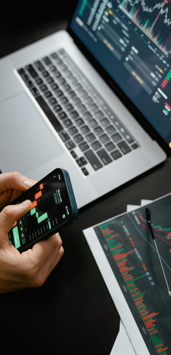 Hands holding phone with financial graph, laptop in view.